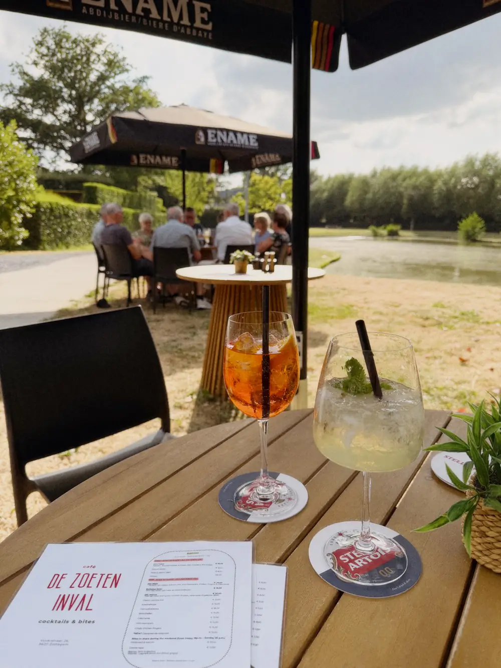 Een Aperol spritz en een gin-tonic op een houten tafel op het terras van De Zoeten Inval, met de vijver, de parasols en gasten op de achtergrond